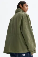 Campera estilo cargo de tejido ripstop, color verde militar, con cuello elevado que incluye capucha oculta. Presenta cierre frontal con solapa rompevientos, cuatro bolsillos delanteros con solapa y botón a presión, puños ajustables y cordón interno en la cintura.