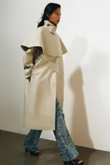 Gabardina larga de corte recto y diseño minimalista en color beige, con cuello solapa, manga larga y cierre frontal con botones. Cuenta con una capa desmontable en el mismo tejido y una solapa decorativa en el pecho.