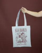 Tote bag de tela en color celeste claro con estampado frontal en color marrón que incluye personajes infantiles en monopatines y la frase 'Sean amables (casi nadie lo está pasando bien)'.