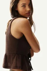 Top corto sin mangas de punto marrón oscuro, con escote redondo y abertura frontal que se cierra con tres lazos anudados. Presenta un ruedo con volado (peplum).