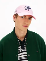 Gorra de béisbol de seis paneles Tommy Hilfiger, color rosado, confeccionada en puro algodón. Presenta un diseño con ojales de ventilación, botón superior y visera curvada con costuras texturizadas. Destaca por el logo de la marca bordado en fuente script azul marino en la parte frontal y lateral.