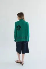Cardigan de punto color marrón con cierre frontal, cuello alto y bolsillos aplicados. Presenta diseño tejido localizado con detalles en violeta y negro en el pecho.