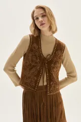 Chaleco de cuero color marrón con bordados decorativos y detalles de tachas metálicas en los bordes. Presenta un cierre frontal mediante lazos finos del mismo material.