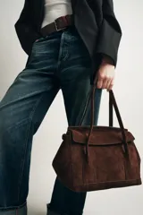 Bolso tipo citybag confeccionado en serraje color marrón oscuro. Presenta una solapa frontal con cierre magnético, asas de hombro integradas y detalles de hebillas metálicas en los laterales para ajuste. Incluye bolsillo interior con cremallera.