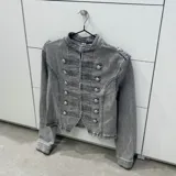 Chaqueta estilo militar de denim gris con efecto acid wash, cuello mao, mangas largas y cierre frontal con múltiples botones metálicos plateados y cordones decorativos.