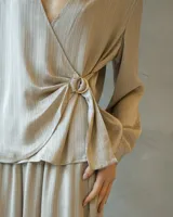 Conjunto de dos piezas en tono arena con textura plisada y acabado satinado. La parte superior es una blusa cruzada de manga larga con escote en V profundo y detalle de hebilla lateral. El pantalón es de corte wide leg con caída fluida.