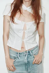 Camisa entallada de color blanco con cuello redondo, manga corta y abertura frontal. Presenta cierre con botones ocultos y detalle de lazadas en el frente.