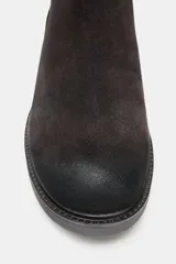 Botín de mujer de serraje color marrón oscuro, estilo motero, con caña media y tres tiras ajustables con hebillas metálicas plateadas. Posee tacón de bloque medio y tirador trasero para facilitar el calce.