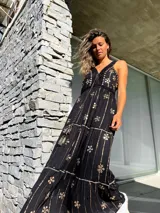 Vestido largo negro con escote en V, breteles finos y estampado floral dorado. Presenta detalles de crochet en el escote y la cintura, y un lazo con borlas en la cintura.