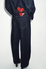 Jeans de tiro alto color azul oscuro, con cinco bolsillos y pernera holgada que se estrecha hacia el tobillo. Presenta pespuntes en color rojo y cierre frontal con cremallera y botón.