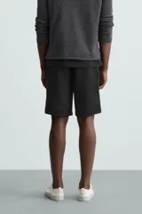 Bermuda negra de corte recto, con cintura elástica ajustable mediante cordón, pliegues frontales y bolsillos de vivo en la espalda. El bajo tiene un dobladillo vuelto.