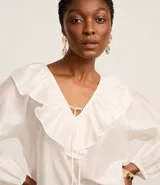 Blusa color blanco confeccionada en voile, de mangas largas con elástico en los puños, cuello en V con volado y lazo con adornos metalizados.