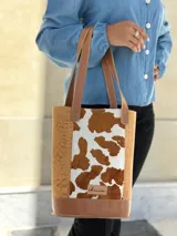 Bolso tipo matera confeccionado en cuero, con diseño combinado que presenta laterales con textura de cocodrilo y panel central con estampado animal print de vaca. Cuenta con asas largas para llevar al hombro y base reforzada.