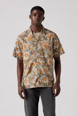 Camisa de manga corta con estampado floral en tonos beige, marrón y azul. Tiene cuello tipo camp y un bolsillo en el pecho.
