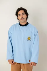 Buzo de cuello redondo color celeste, con bolsillo frontal bordado que presenta el sol de la bandera uruguaya, cuatro estrellas y la frase 'Heroicos sabremos cumplir'.