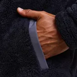 Canguro de sherpa con cierre frontal completo, cuello alto y bolsillo en el pecho con cierre. Presenta una combinación de texturas en color negro y gris oscuro, con diseño funcional y térmico.