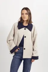 Chaqueta corta de color beige con cuello y puños vueltos en contraste de color azul marino, cierre frontal de botones oscuros y bolsillos ribeteados.