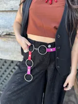 Cinturón de cuero con herrajes metálicos circulares grandes y tiras de cuero en tonos rosa y fucsia que cuelgan con aros metálicos adicionales.
