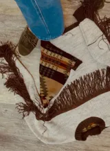 Buzo de punto con diseño western, que combina cuerpo y mangas tejidas en color crudo con un panel central de paño estampado con trama geométrica estilo Pendleton. Presenta flecos de gamuza marrón en el frente y cuello redondo tejido en color marrón.