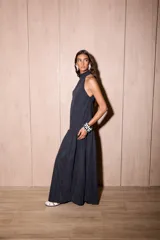 Vestido largo negro, holgado, con escote halter y espalda baja. Presenta un detalle de volado en el cuello y un maxi volado en la cadera. El material es tela Cupro, con apariencia similar a la gamuza.