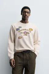 Sudadera de cuello redondo y manga larga en color beige. Presenta un diseño con texto bordado y múltiples parches decorativos en el frente, con terminaciones en rib en puños y cintura.