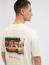 Camiseta de manga corta y cuello redondo, color blanco antiguo (off-white), con un estampado gráfico grande en la espalda que simula una fotografía urbana nocturna con un taxi amarillo y texto en tipografía cursiva y recta.