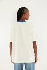 Camiseta blanca de algodón con cuello redondo azul, manga corta y gran estampado frontal con la palabra BRASIL, la bandera de Brasil y un pájaro verde en vuelo.