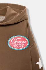 Sudadera marrón con capucha ajustable con cordones, manga larga con puños y bajo elástico. Presenta un estampado ovalado en el pecho con el texto "Texas USA" en rosa y azul, y un diseño de estrellas blancas en las mangas. El bolsillo canguro frontal tiene detalles de tachas y bordes deshilachados.