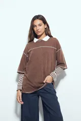 Remera de manga corta color marrón con efecto desgastado, corte oversize y costuras a la vista en color blanco. Presenta cuello redondo con cuello polo blanco superpuesto y mangas largas a rayas horizontales marrones y blancas por debajo.