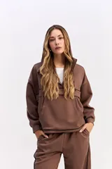 Hoodie de algodón color marrón con capucha, cierre frontal parcial y detalle de costuras horizontales en contraste negro. Presenta diseño holgado, mangas largas con puños elásticos y cordones ajustables en la capucha.