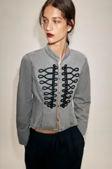 Chaqueta de denim gris con cuello subido y manga larga. Presenta un diseño entallado con bajo en evasé y un llamativo bordado frontal de alamares en color negro. Cierre frontal con corchetes ocultos.