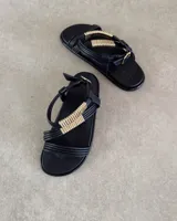 Sandalias planas de color negro con tiras finas y detalle decorativo de tubos metálicos dorados en el empeine. Cuentan con cierre de hebilla ajustable en el tobillo.