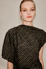 Vestido midi de corte asimétrico con escote barco, una manga corta y un hombro descubierto. Presenta un diseño drapeado en el lateral y estampado de lunares dorados sobre fondo negro.