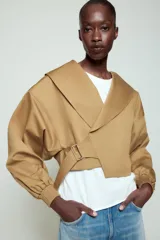 Chaqueta corta de Zara confeccionada en mezcla de algodón, con cuello solapa, manga larga con hombro caído y puños elásticos. Presenta un diseño cruzado con cierre de hebilla metálica lateral y forro interior a tono.