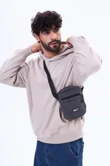 Morral de diseño compacto en color gris oscuro, con cierre principal y bolsillo frontal con cierre. Incluye una etiqueta decorativa con texto en el frente y correa ajustable para llevar al hombro o cruzado.