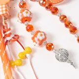 Llavero decorativo en tonos naranja, compuesto por una cuerda trenzada, un mosquetón en forma de corazón y múltiples dijes colgantes que incluyen cuentas de cristal, figuras de flores, un dije de corazón de cuero sintético y una esfera brillante.