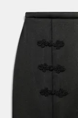 Falda midi de tiro medio y corte recto, confeccionada en tejido satinado color negro. Presenta detalle de alamares decorativos a tono en el frente y cierre con cremallera en la espalda.