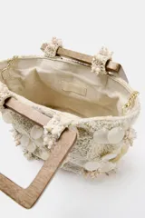 Bolso de mano formato mini confeccionado en tejido texturizado color crema. Presenta apliques de nácar y bordados florales en relieve, con doble asa rígida de madera y cadena bandolera extraíble.