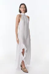 Vestido largo de lino color blanco, con cuello halter alto y diseño sin mangas. Presenta un tajo frontal decorado con un amplio volante u olán en el borde.