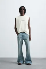 Camiseta sin mangas (manga sisa) de punto color crema, corte relaxed fit, con cuello redondo y acabados irregulares en las sisas. Presenta un texto estampado en el pecho que dice "SERIAL N. 8763/ 7.3Z".