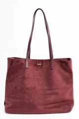 Bolso tipo tote de gran tamaño en color bordó, confeccionado en material con textura similar a la gamuza. Cuenta con doble asa larga para llevar al hombro y una pequeña placa metálica decorativa en el frente.
