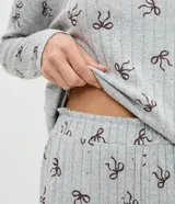 Pijama de dos piezas confeccionado en tejido de punto, compuesto por una blusa de manga larga con cuello redondo y puños ajustados, y un pantalón recto con cintura elástica. Presenta un estampado continuo de moñitas sobre fondo gris.
