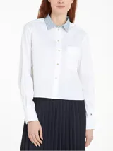 Camisa blanca de algodón orgánico, nailon y elastano, con cuello de rayas celestes y cierre con botones dorados. Tiene corte boxy, manga larga, bolsillo de parche en el pecho y logo de Tommy Hilfiger bordado en el puño.