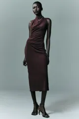 Vestido midi de color negro, con cuello subido y diseño sin mangas. Presenta un detalle de drapeado lateral en la parte delantera y cierre de cremallera oculta en la espalda.