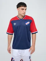 Remera de fútbol Umbro del Club Nacional de Football, de diseño tricolor con cuerpo azul marino, mangas y canesú rojos, y cuello en V con detalles en blanco y azul. Incluye el logo de Umbro en el pecho derecho y el escudo del club en el izquierdo.