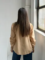 Blusa de color tostado con cuello en V, cierre frontal con botones y mangas largas con puños elásticos. Presenta un diseño de corte recto con bolsillos laterales integrados.