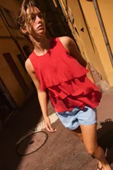 Top de cuello redondo y manga sisa, confeccionado en tejido con acabado satinado. Presenta volantes en cascada y un diseño de bajo asimétrico, con cierre en la espalda mediante pequeña abertura y botón.