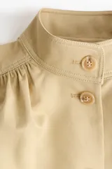 Campera corta de sarga color beige, con cuello alto, cierre frontal de botones, mangas largas abullonadas con puños ajustados y bolsillos laterales ribeteados.