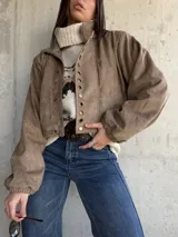 Campera bomber de gamuza color beige, con cierre frontal y detalle de ojales metálicos decorativos en la solapa. Presenta mangas largas con puños elásticos y un corte holgado tipo oversize.