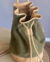 Mochila tipo bucket de gamuza marrón, con detalles de cuero liso en el tono, bolsillo frontal con cierre y cordón ajustable con tiras.
